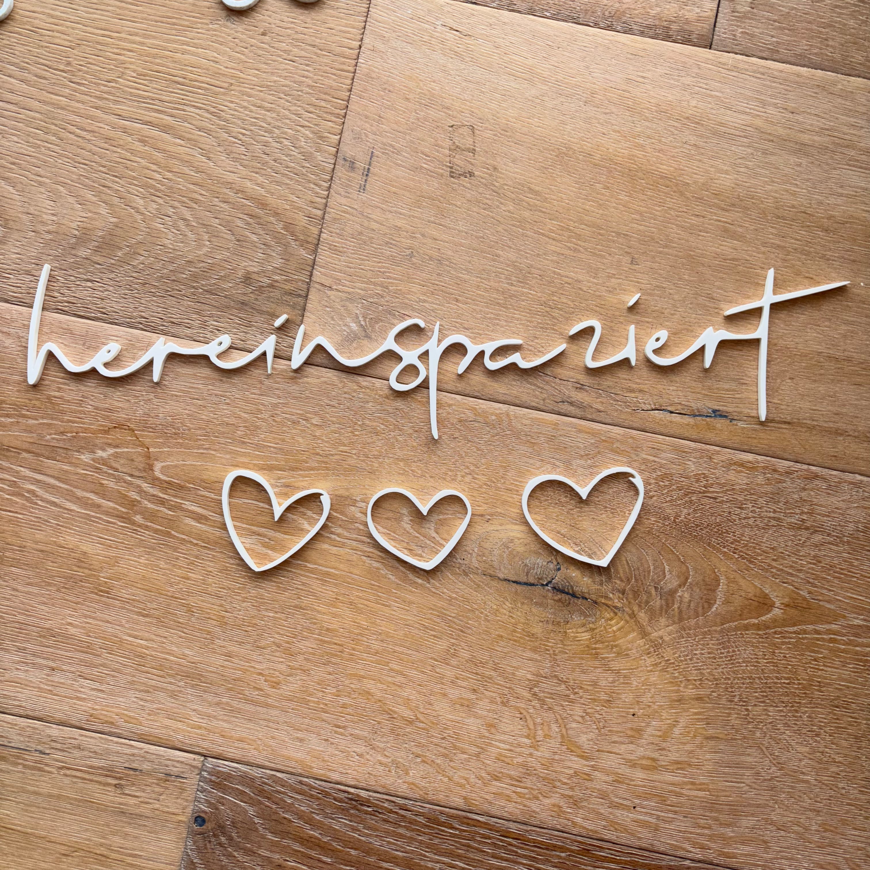 Weißes 3D-Wandschild „hereinspaziert“ mit drei kleinen Herzen, flach auf einem Holzfußboden arrangiert.