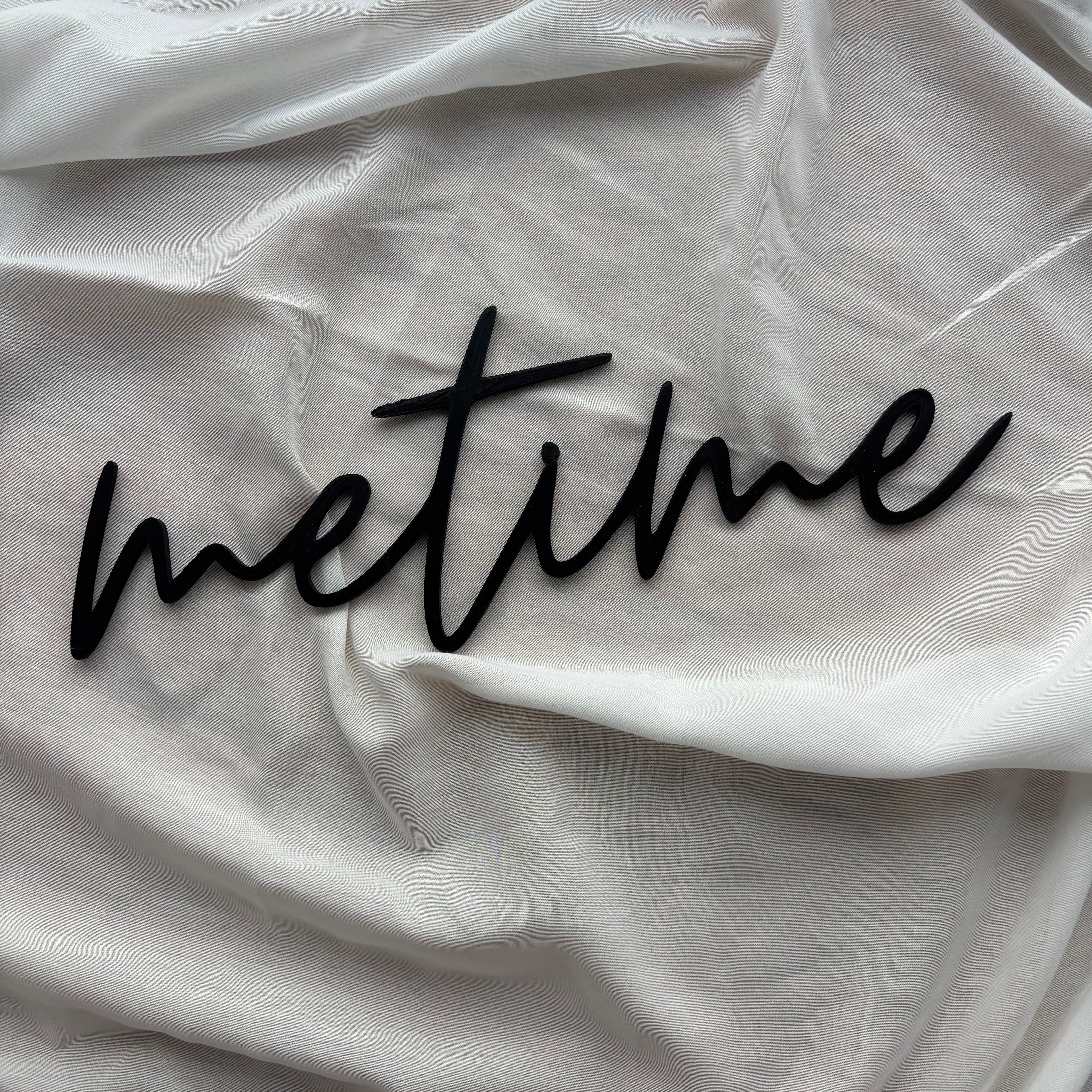 Schwarzer 3D-Schriftzug „metime“ aus PLA, dekorativ auf grauem Stoff drapiert.
